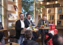 en panelsamtale med tre menn som sitter ved høye bord i et bibliotek