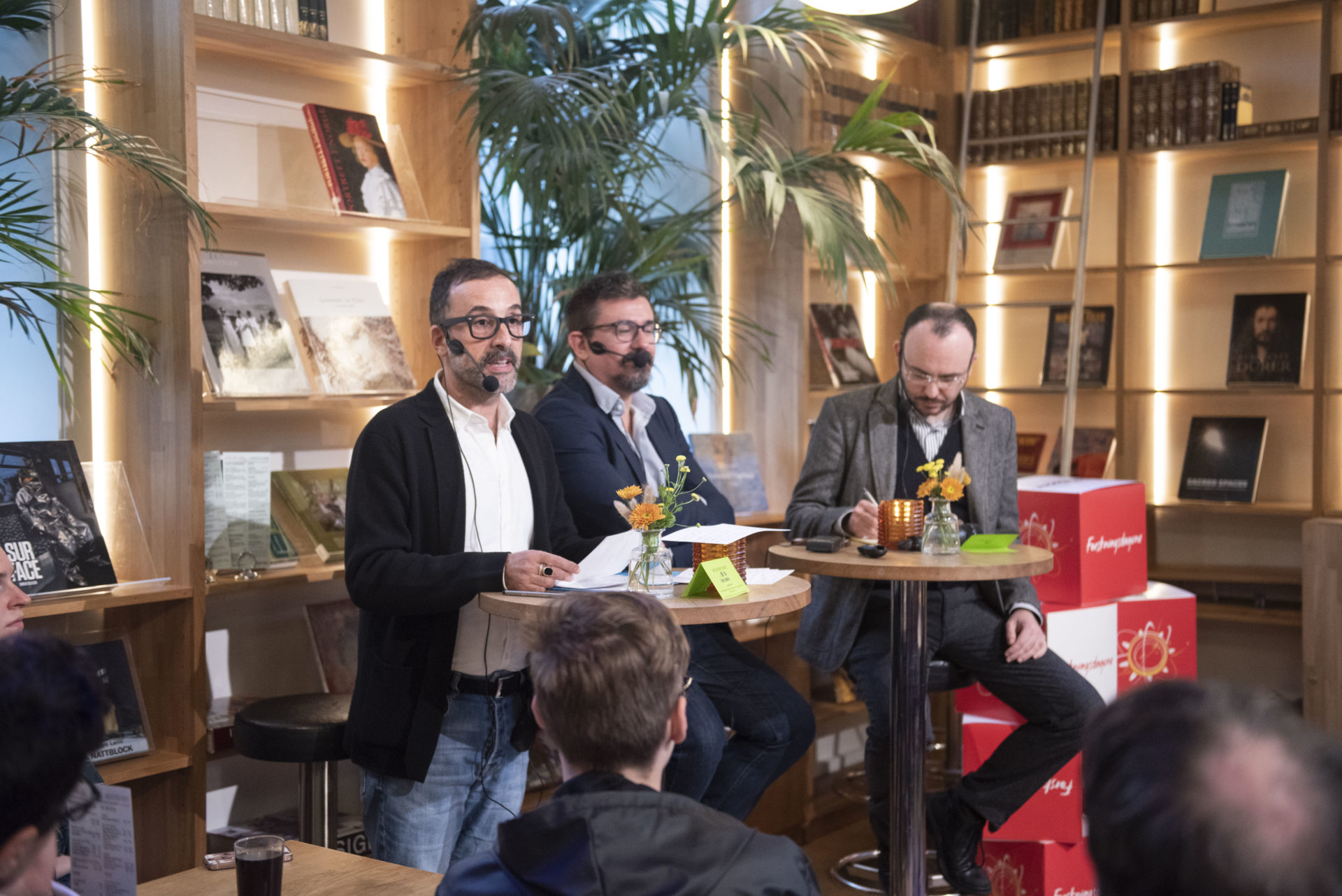 en panelsamtale med tre menn som sitter ved høye bord i et bibliotek