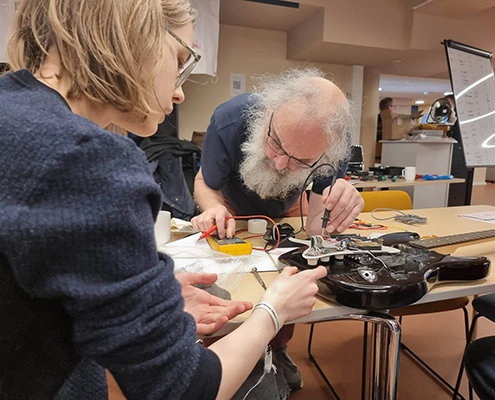 Mann og dame holder på å skru på en el-gitar på et bord fullt av verktøy