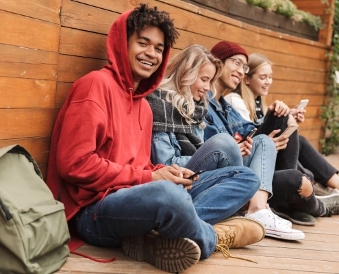 Ungdommer som sitter lent inntil en vegg. De sitter med hver sin smartelefon og stirrer på den. En gutt smiler mot kamera.