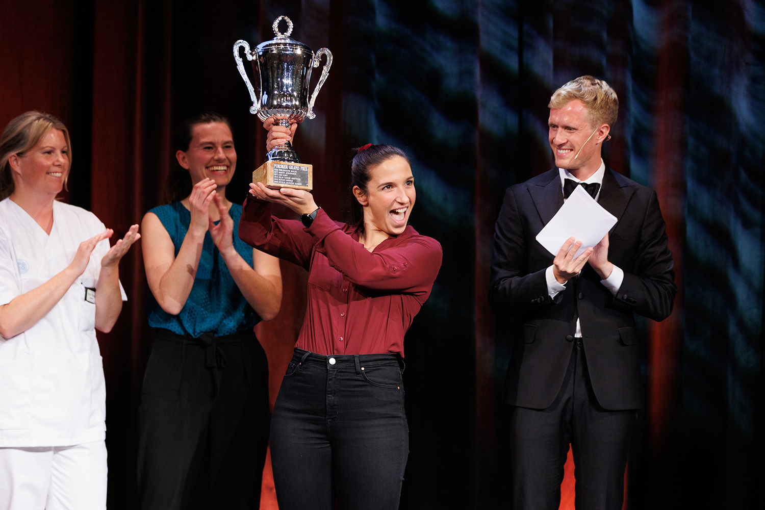 Vinner av Forsker Grand Prix Josepine Salice holder opp pokalen hun vant, mens de andre deltakerne og programleder applauderer.