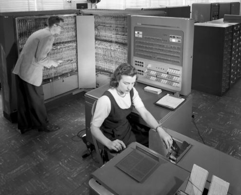 Sort-hvitt bilde fra 50-tallet av en datamaskin, som opptar hele rommet og to teknikere.